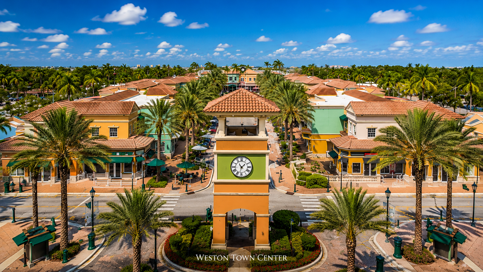 Aerial view of Weston Town Center showing a central clock tower surrounded by pastel storefronts and palm trees under a blue sky.